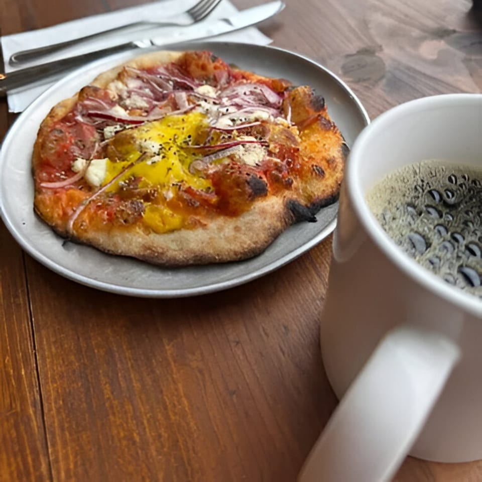 Cafe Enza's breakfast pizza with egg, bacon, and onions, served with a cup of coffee.