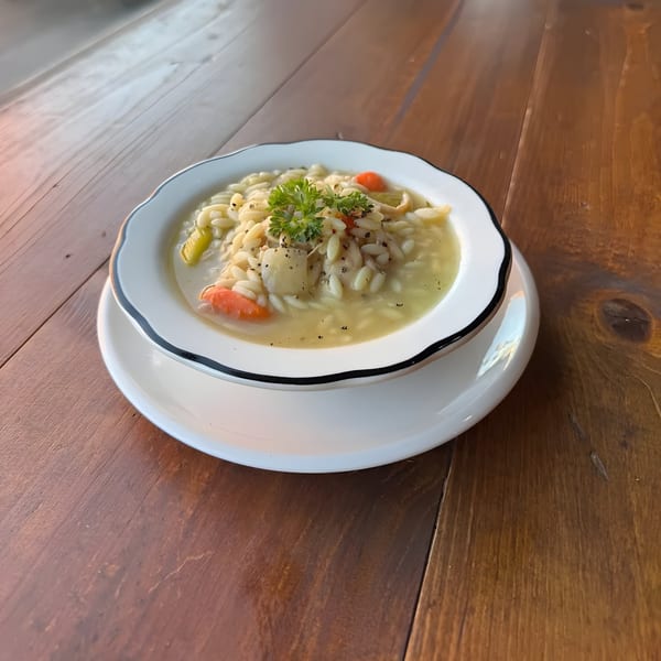 A bowl of Cafe Enza's chicken orzo soup with carrots, celery, and parsley on a wooden table.