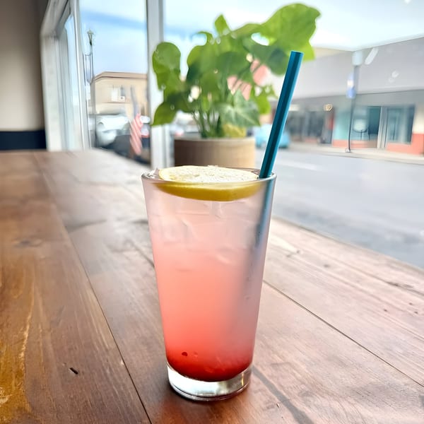 Pink lemonade with a lemon slice and blue straw on a wooden table at Cafe Enza.