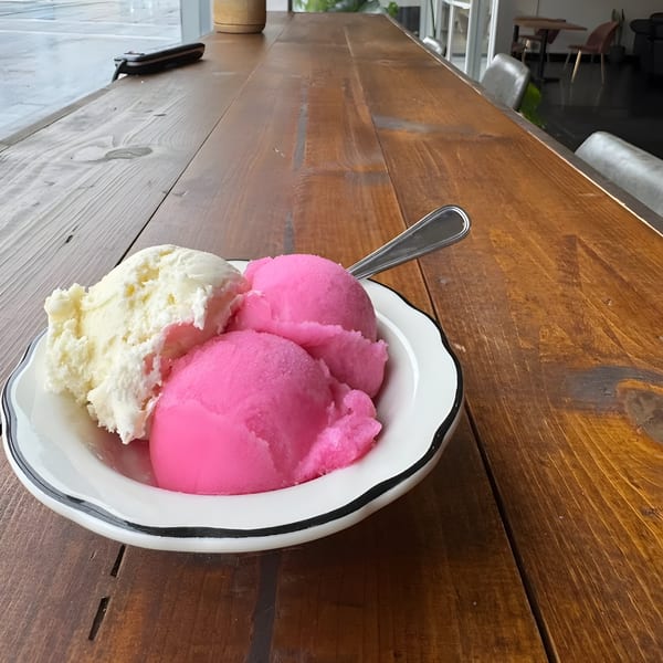 A white bowl with scoops of pink and white ice cream sits on a long wooden table.